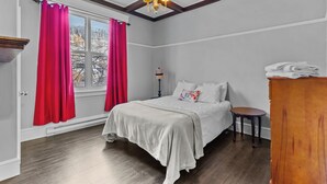 Una camera da letto con un letto, un comodino, una cassettiera in legno, una finestra con tende rosse e un lampadario.