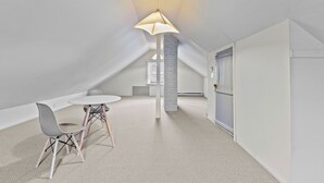 A spacious attic room with a high ceiling, a central light fixture, a round table with chairs, and a door on the right.