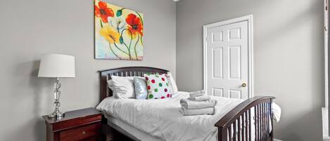 Un dormitorio con una cama, una mesita de noche, una lámpara, una puerta y un cuadro en la pared.