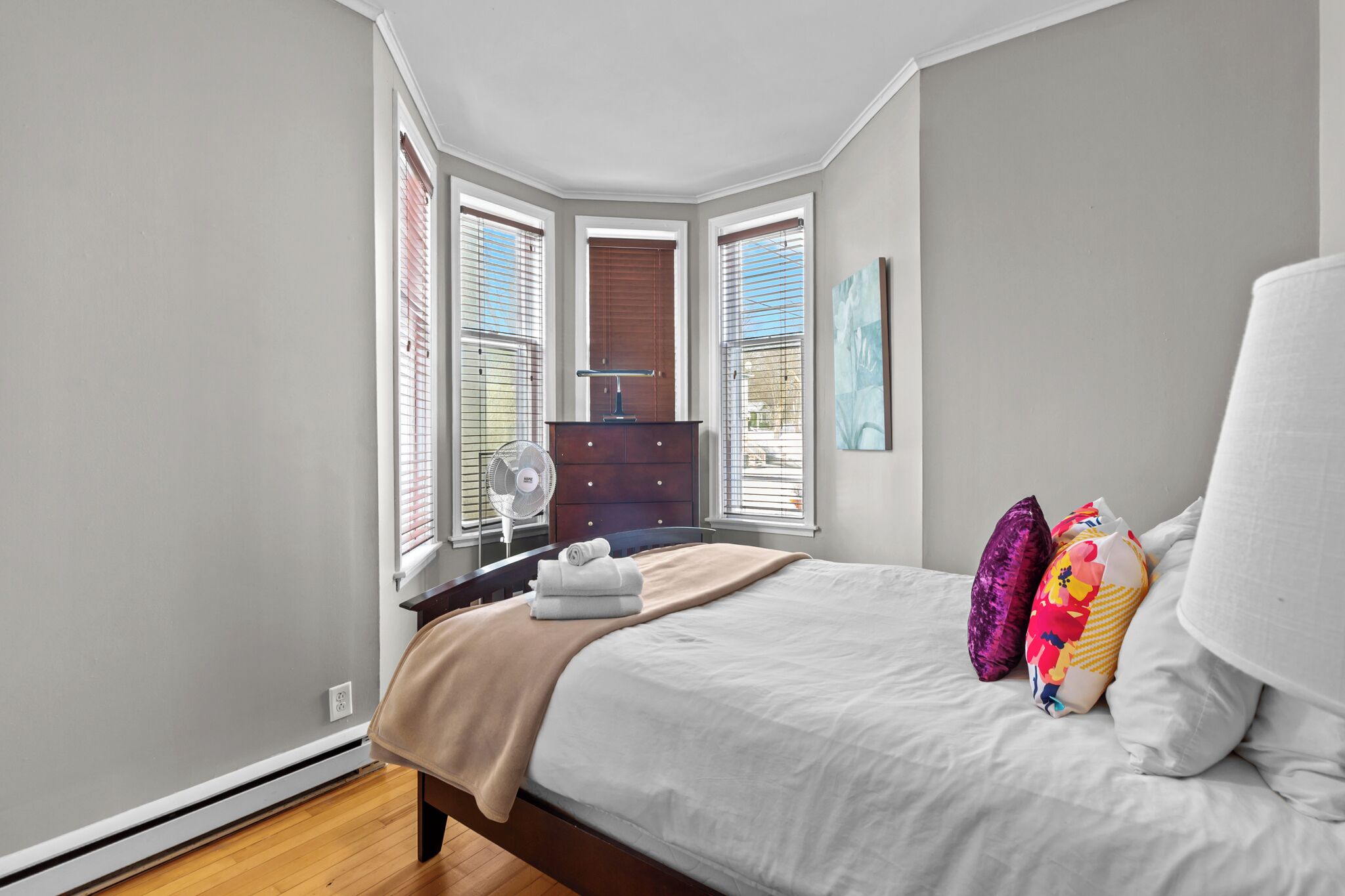 Une chambre à coucher avec un lit, une commode, un ventilateur et une fenêtre avec des stores.