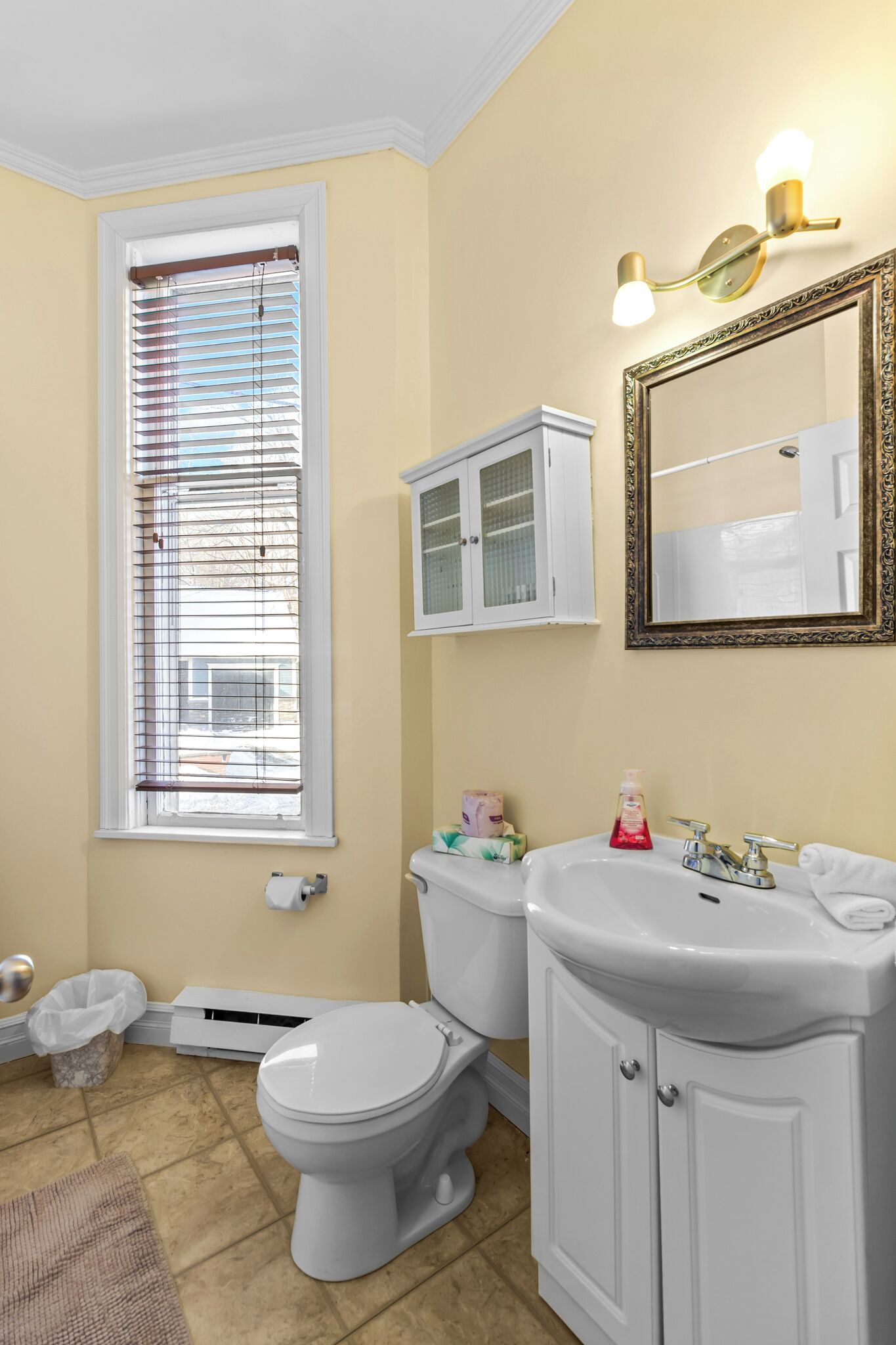 Une salle de bain équipée d’un WC, d’un lavabo, d’un miroir, d’un meuble et d’une fenêtre avec des stores.