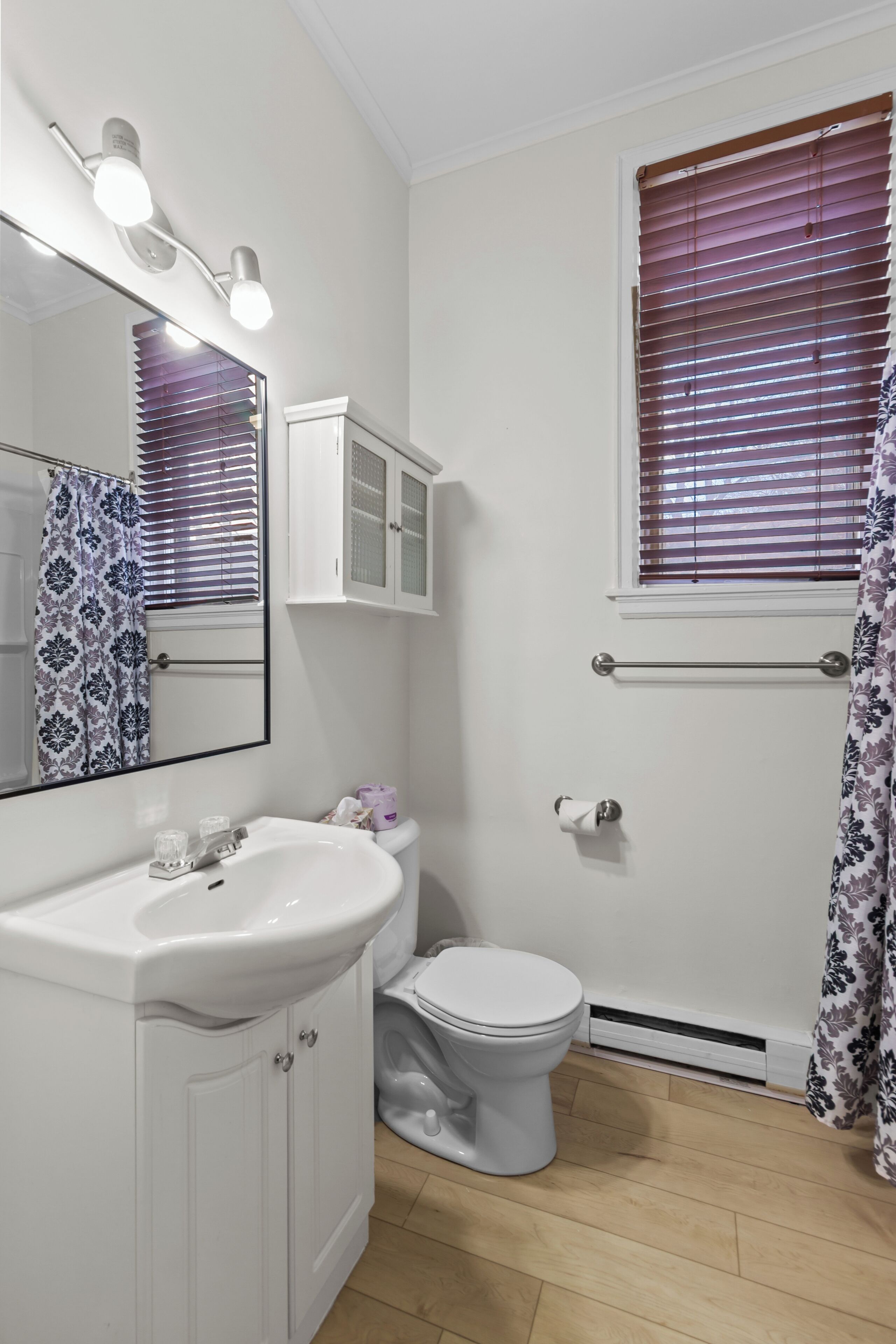 Une salle de bain avec un lavabo blanc, un miroir et des toilettes. Il y a une fenêtre avec des stores et un rideau de douche.