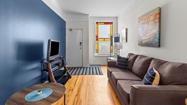 A modern living room with a brown sofa, a television, a small table with a plant, and a window with a stained glass design. - Charming 2BR Wi-Fi Off-Street Parking (St. John's)
