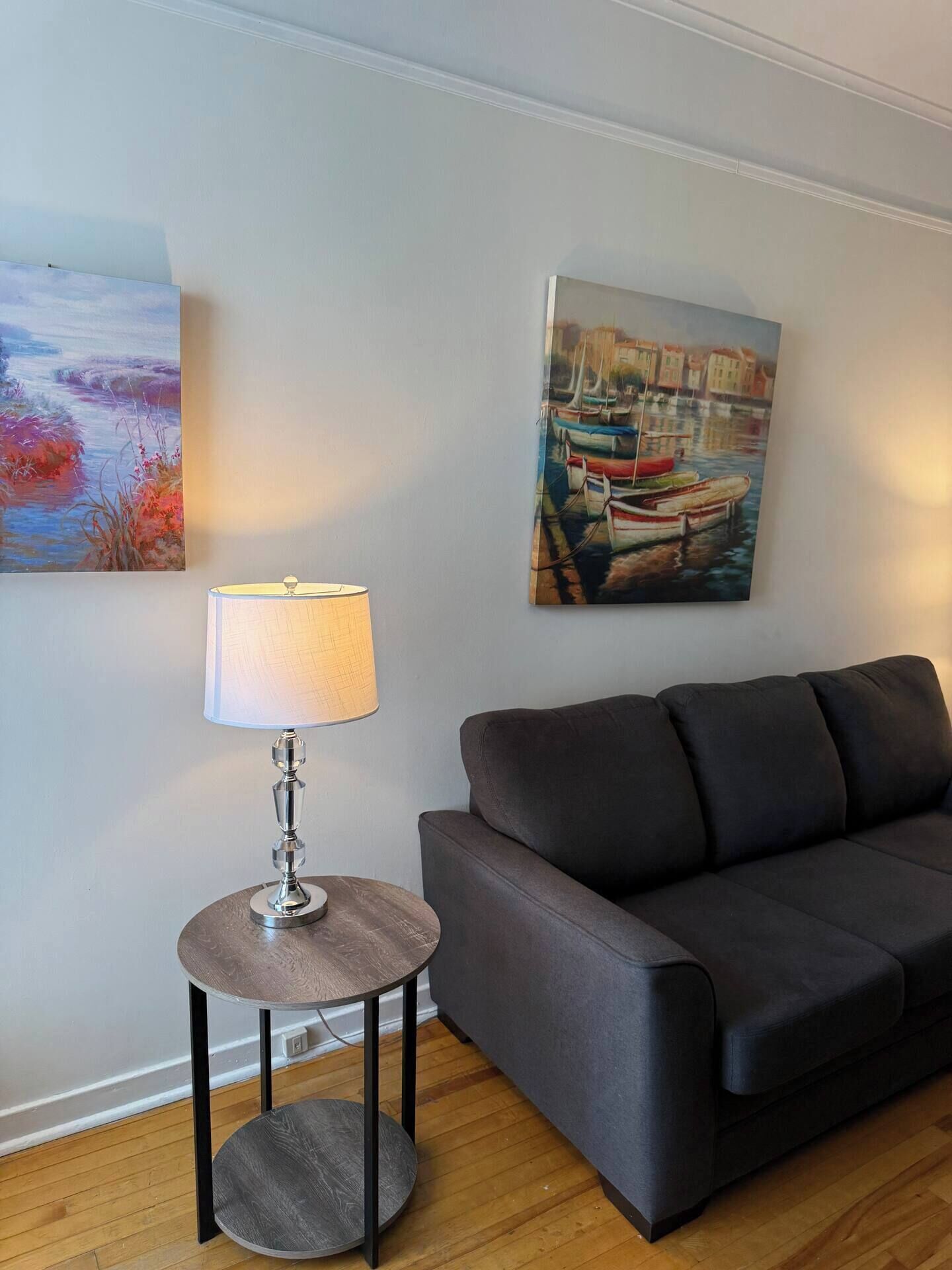 A living room with a dark grey sofa, a round wooden side table with a lamp, and two paintings on the wall.