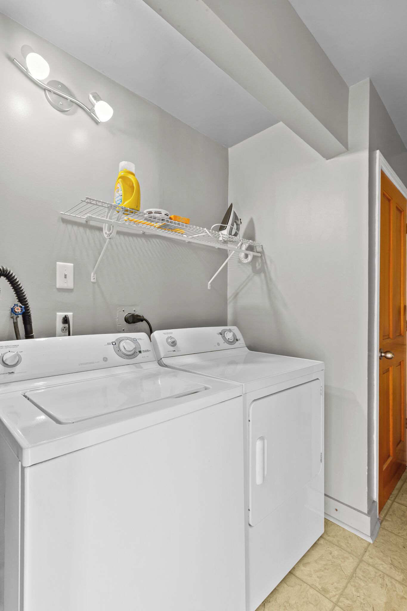 A laundry area with two white front-loading washers and dryers, a metal drying rack with a yellow detergent bottle, and a wall-mounted light fixture.