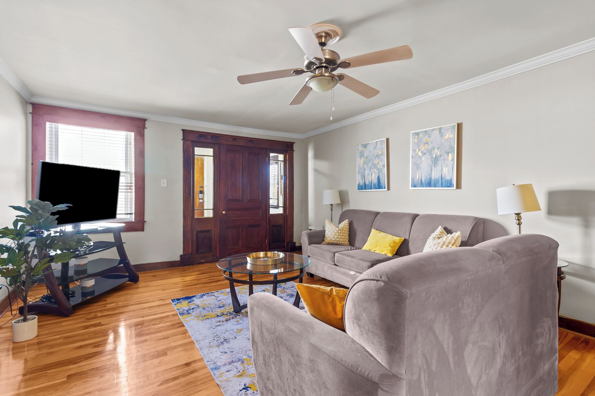 A living room with a grey sofa, a coffee table, a TV on a stand, a ceiling fan, and artwork on the walls.