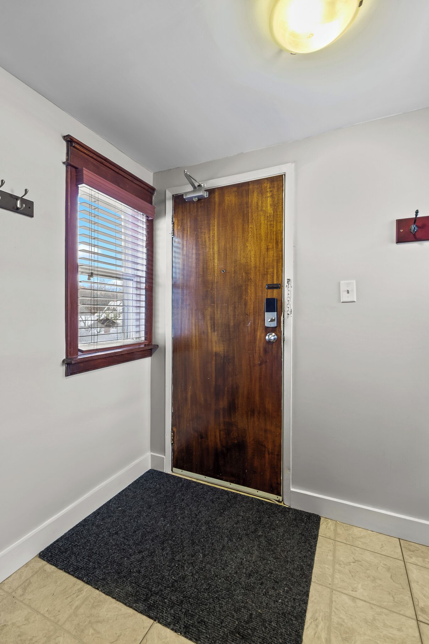 A room with a wooden door, a window with blinds, a carpeted entrance, and a light fixture on the ceiling.