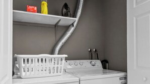Une buanderie équipée d’un lave-linge et d’un sèche-linge à chargement frontal, d’un panier à linge blanc, d’étagères contenant des produits de lessive et d’un tuyau d’aération.