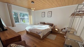 A bedroom with a wooden floor, a bed with a white and blue patterned bedspread, a wicker chair, a desk, a small table, and a hanging rack. - Modern vacation apartment in Marbach am Neckar in the immediate vicinity of the Schillerhöhe (Marbach am Neckar)