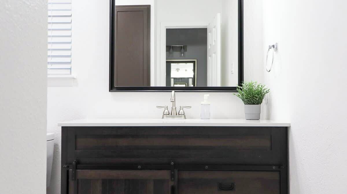 Un baño con un mueble de tocador de madera oscura, una encimera blanca, un espejo, una planta en maceta y un aro para toallas en la pared.