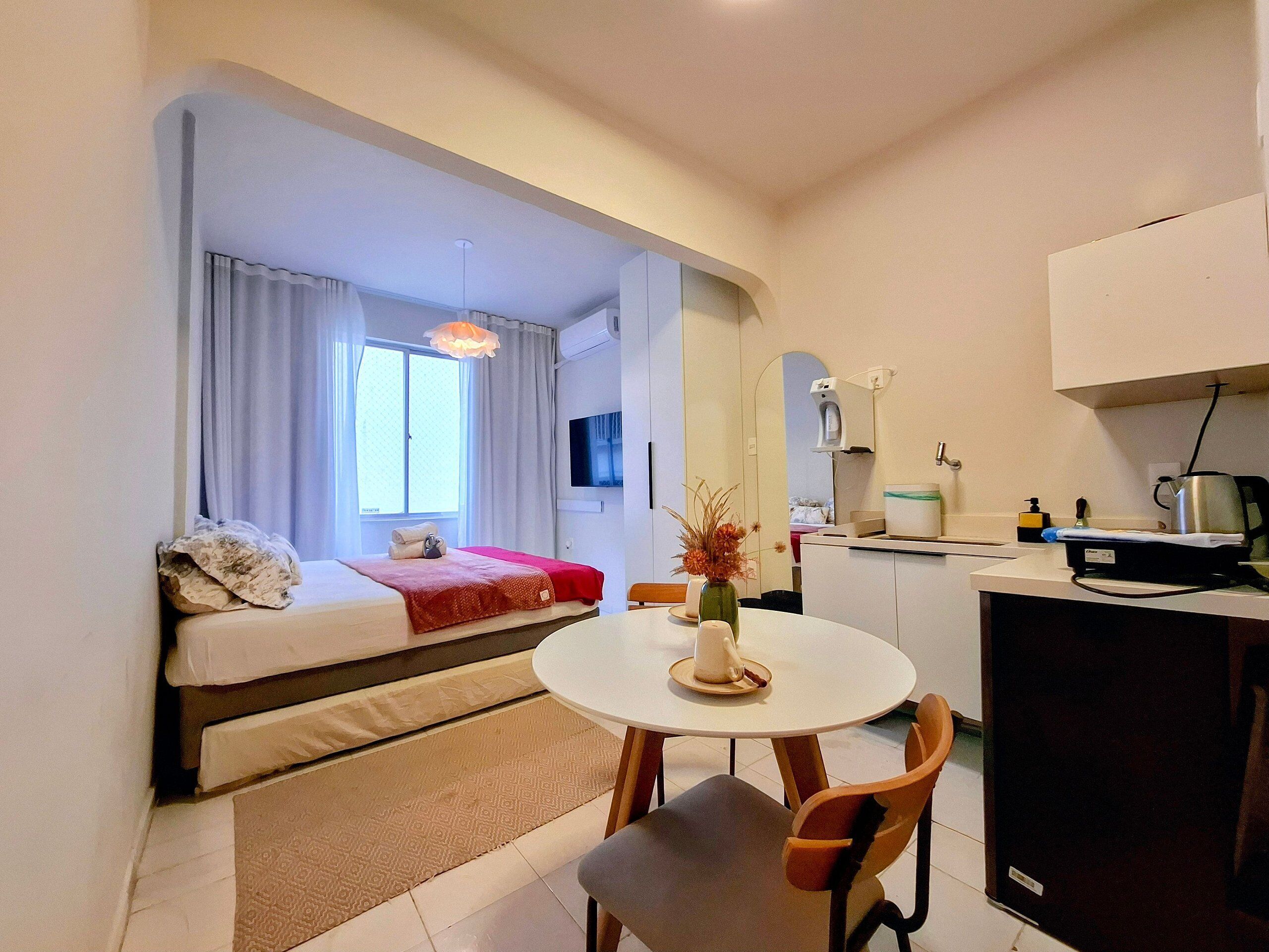 Een compacte hotelkamer met een bed, een eettafeltje, een stoel, een kitchenette en een badkamer zichtbaar via een boog.