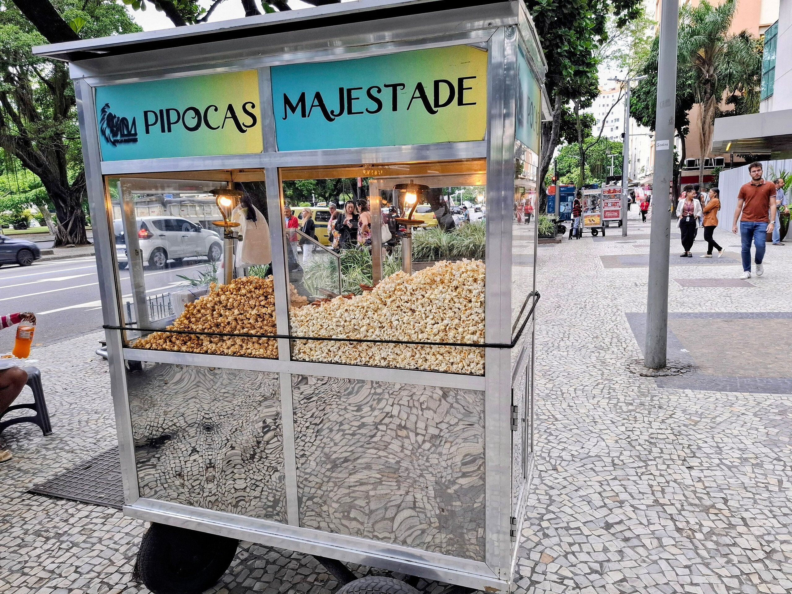 Un carretto di venditore di popcorn con la scritta 'PIPOCAS MAJESTADE', situato su una strada acciottolata con pedoni e veicoli sullo sfondo.