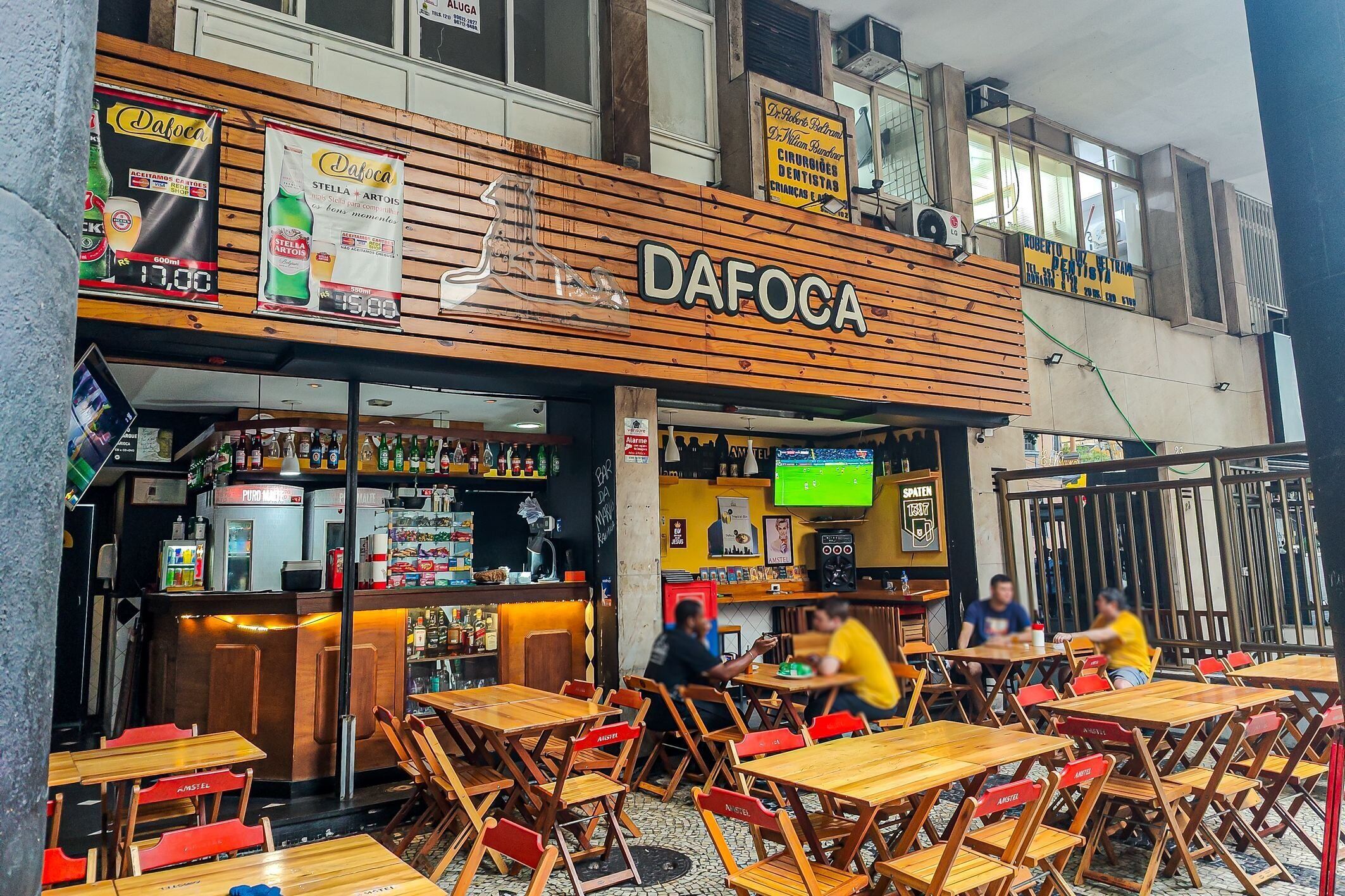 Un'area salotto esterna con tavoli e sedie di legno, un bar con bancone e un cartello con la scritta 'DAFOCA' sopra l'ingresso.