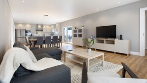 Uma sala de estar moderna com sofá, mesa de centro e televisão. Há uma área de jantar com cadeiras e mesa, e uma cozinha ao fundo.