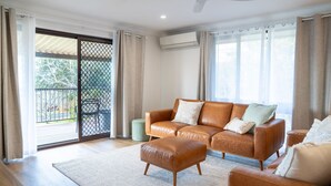 A spacious living room with a leather sofa, a matching ottoman, and a large window with curtains. - Perfectly positioned between Woolgoolga’s beautiful beaches (Woolgoolga)