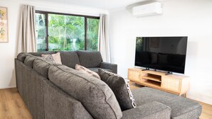 A modern living room with a grey sectional sofa, a flat-screen TV on a wooden stand, and a large window with curtains. - Perfectly positioned between Woolgoolga’s beautiful beaches (Woolgoolga)