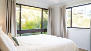 A hotel room with a bed, curtains, and large windows offering a view of greenery and buildings. - Perfectly positioned between Woolgoolga’s beautiful beaches (Woolgoolga)