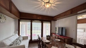 Ein gemütliches Zimmer mit Esstisch für vier Personen, einer Couch, einem Fernseher und einem großen Fenster mit Vorhängen.