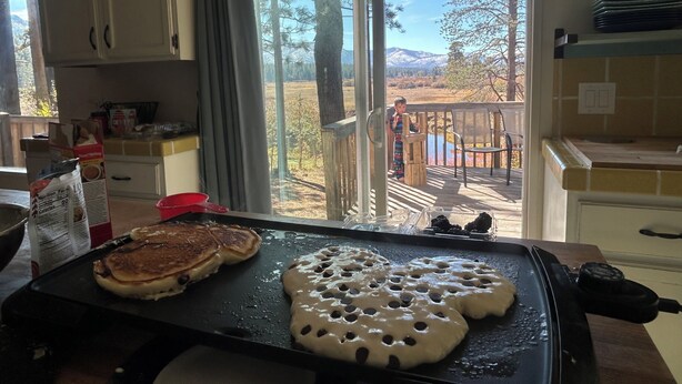 Una cocina con una plancha preparando panqueques y gofres, un cartón de leche, y vistas a una terraza con un paisaje de montaña.