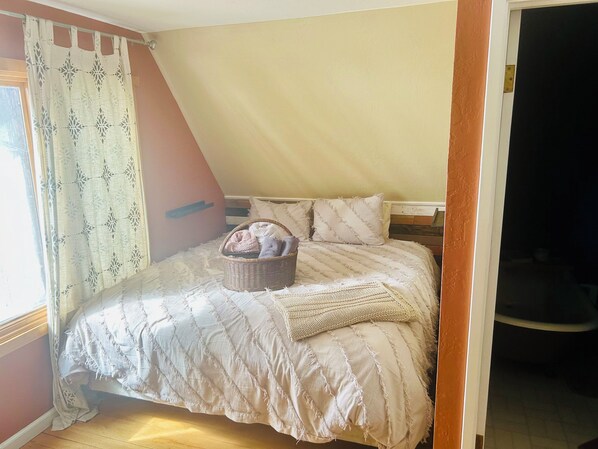 A bedroom with a bed, a basket of towels, and a window with patterned curtains.