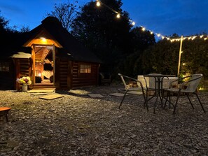 A cozy log cabin with a welcoming entrance, a small patio area with wicker chairs, and string lights creating a warm ambiance. - Hobbit Haven Hot Tub and Countryside Bliss (Gaerwen)