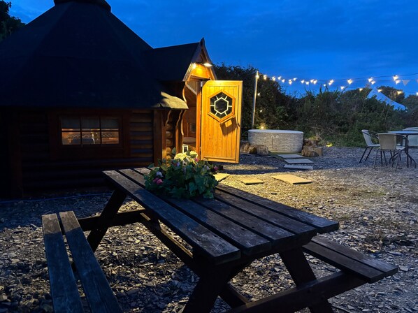 Une cabane en bois avec une porte hexagonale, une table de pique-nique et des guirlandes lumineuses.