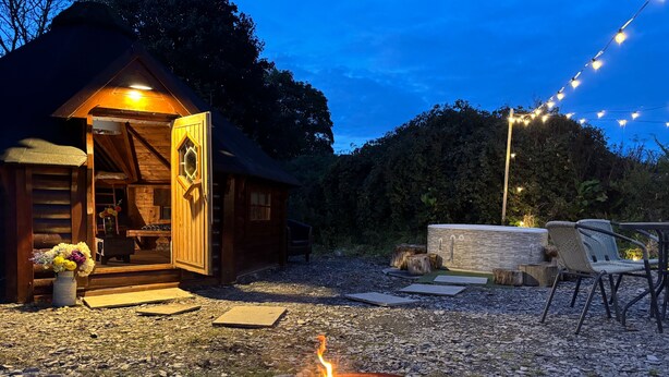 A rustic wooden cabin with a fire pit in the foreground, surrounded by a gravel path and outdoor furniture. - Hobbit Haven Hot Tub and Countryside Bliss (Gaerwen)