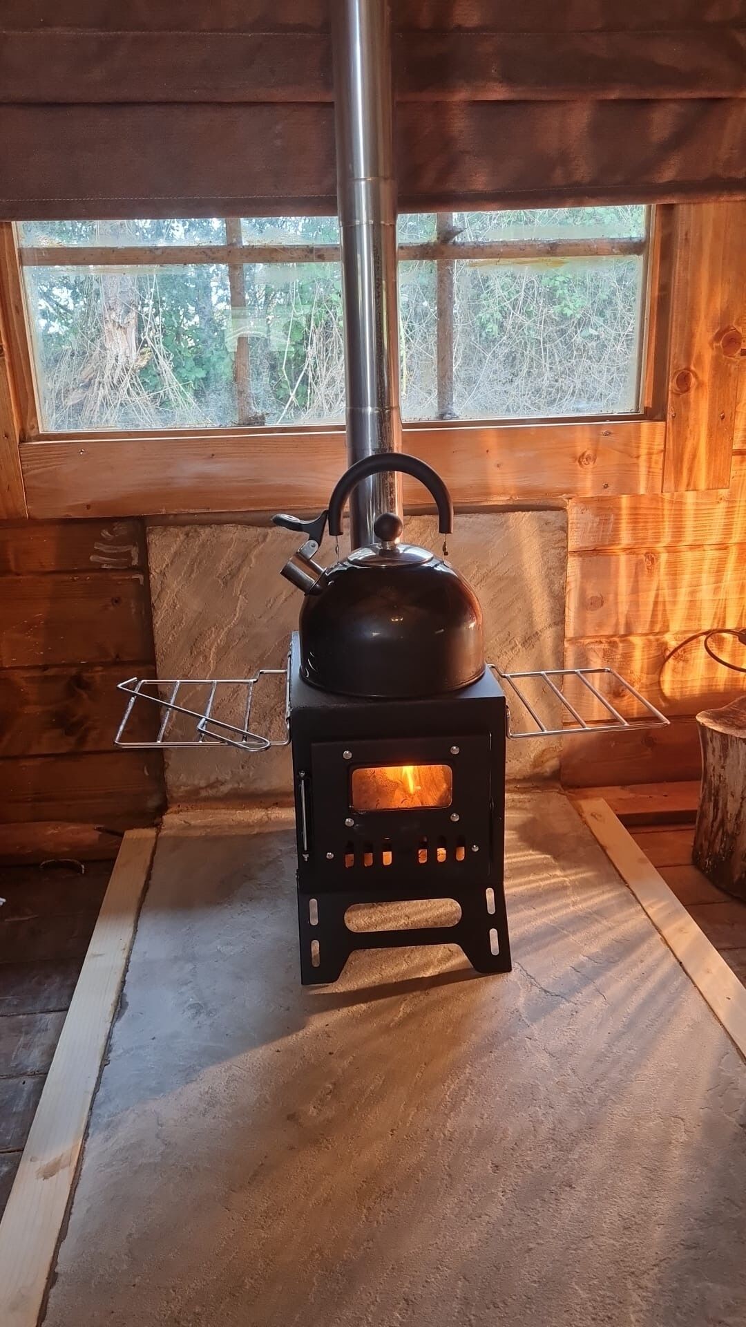 En brændeovn med en kedel ovenpå, placeret i en træhytte med et vindue der kigger ud over en skov.