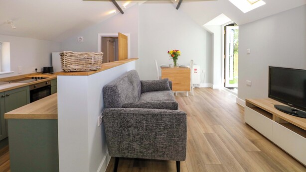 A modern kitchen with a built-in island, a grey sofa, and a TV in a living area with wooden flooring. - The Old Parlour (Southampton)