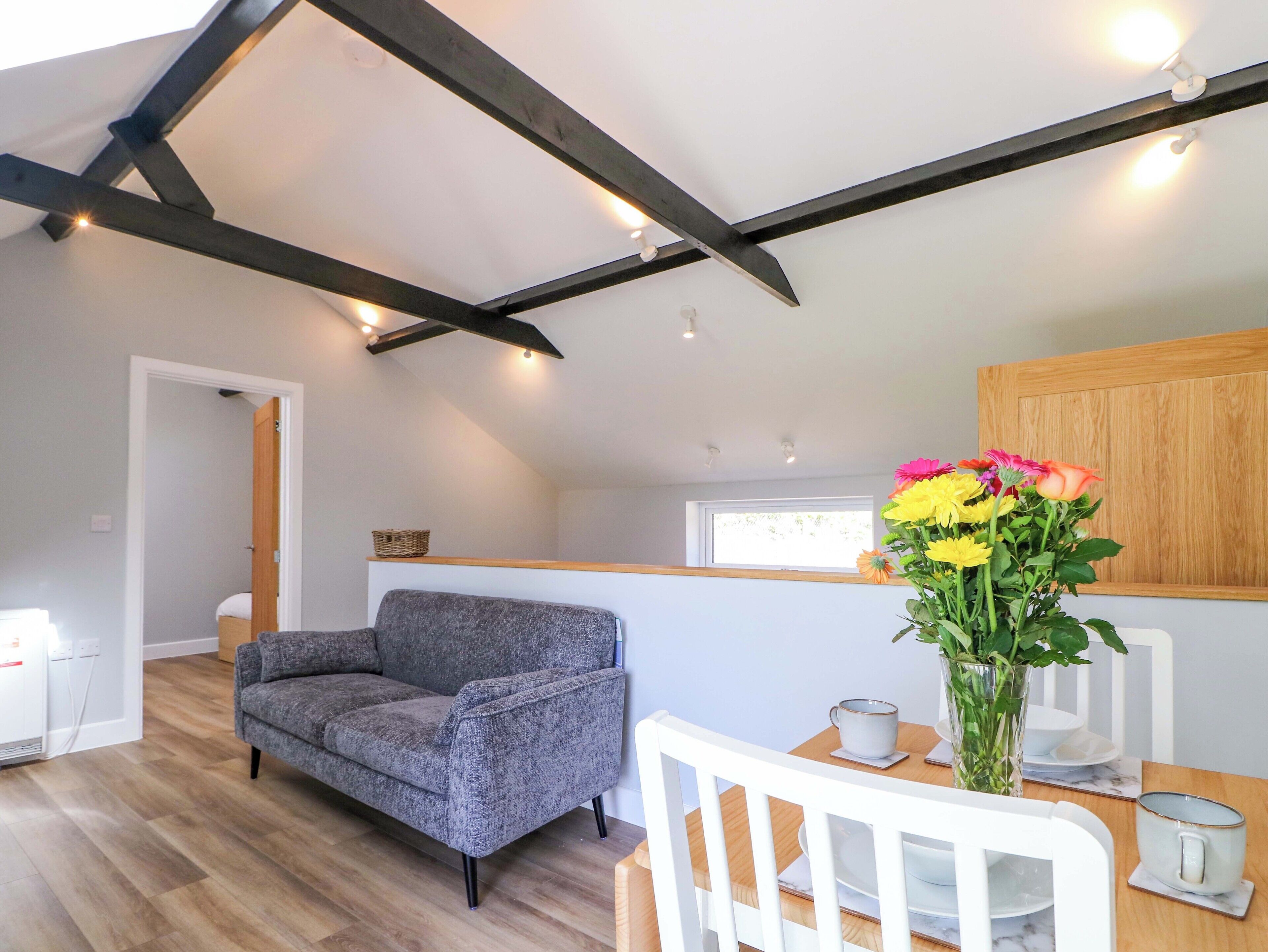A modern loft bedroom with exposed beams, a gray sofa, a dining table with white chairs, and a vase of flowers.