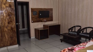 Una camera d'albergo con una porta in legno, una TV fissata al muro, una scrivania, due sedie e un letto con copripiumino floreale.