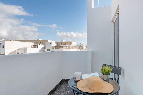 Ein Balkon mit weißer Wand, einem kleinen Tisch, einem Hut, einer Tasse und einem Buch sowie einer Topfpflanze.