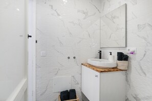 Casa de banho com azulejos de mármore branco, bancada de madeira, lavatório branco, espelho e cesto com toalhas.