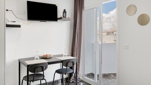 A modern room with a flat-screen TV, a dining table with chairs, a sliding glass door to a balcony, and a small shelf with decorative items.