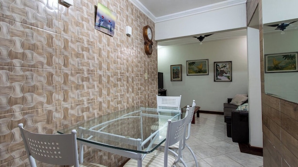 A dining area with a glass table and white chairs, a patterned wall, and a clock. - Apto Pet Friendly c Churrasqueira - Praia do Morro (Guarapari)