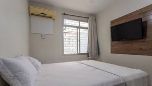 A single bed with white bedding, a window with curtains, an air conditioning unit, and a mounted television on the wall. - Apto Pet Friendly c Churrasqueira - Praia do Morro (Guarapari)