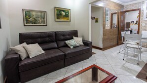 A living room with a dark brown leather sofa, two framed paintings, a glass coffee table, and a kitchen area visible through an open doorway. - Apto Pet Friendly c Churrasqueira - Praia do Morro (Guarapari)