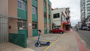 A narrow urban street with a blue electric scooter parked on the sidewalk, a red car parked on the street, and a building with a green and beige facade. - Apto Pet Friendly c Churrasqueira - Praia do Morro (Guarapari)