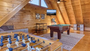 A cozy, wooden-floored room with a foosball table, a pool table, and a television mounted on the sloped ceiling. - Cozy Cabin with Amazing Views! Sleeps 8! Private Hot Tub! Resort Pools/Mini Golf (Sevierville)