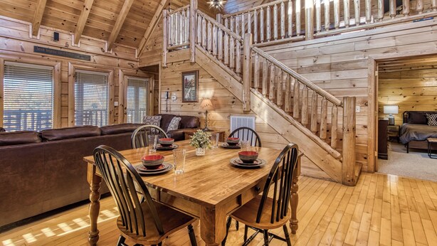 A wooden cabin interior with a dining area, a staircase, and a living room with a sofa and a fireplace. - Cozy Cabin with Amazing Views! Sleeps 8! Private Hot Tub! Resort Pools/Mini Golf (Sevierville)