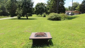 A square outdoor grill with a glass top, surrounded by a grassy field with trees and a building in the background.
