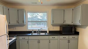 A kitchen with light gray cabinets, a black countertop, a sink, a stove, a microwave, and a window with a view of trees.