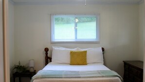A bedroom with a bed, a window with blinds, a bedside table with a lamp, and a dresser.