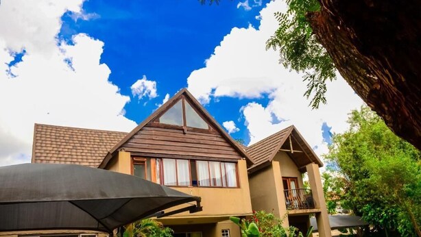 A two-story house with a brown roof and beige walls, surrounded by lush greenery and a clear blue sky. - Novus Guest House (Pretoria)