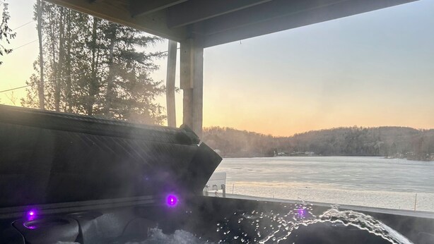 Un jacuzzi con chorros de agua, vista a un lago congelado y una colina boscosa al fondo.