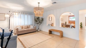 Una sala de estar moderna con un sofá, un banco, una alfombra grande y un área de comedor visible a través de un arco.