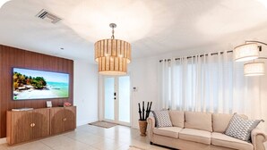 Una sala de estar moderna con un televisor de pantalla plana, un sofá color beige, un mueble de entretenimiento de madera y un ventanal con cortinas translúcidas.