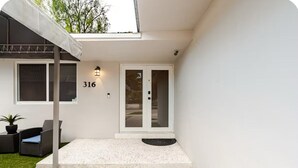 Una casa moderna con fachada blanca, el número '316' en la pared, un porche cubierto con una silla y una mesita, y una puerta con paneles de vidrio.