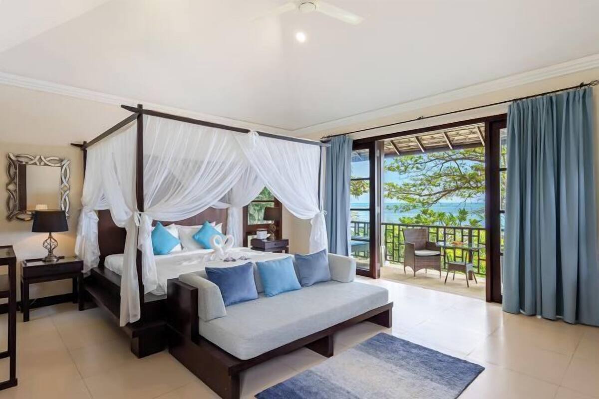 A bedroom with a four-poster bed, a sofa, a nightstand, and a view of the outdoors.