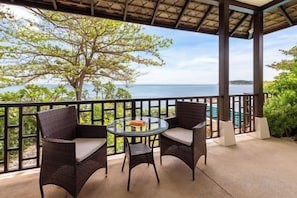 A covered outdoor seating area with wicker furniture and a table, overlooking a scenic view of the sea and a distant island.